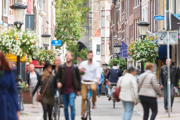 A softly focused scene of people walking and cycling down a charming European street lined with historic buildings, street lamps, and hanging flower baskets, conveying a sense of daily life in the city.